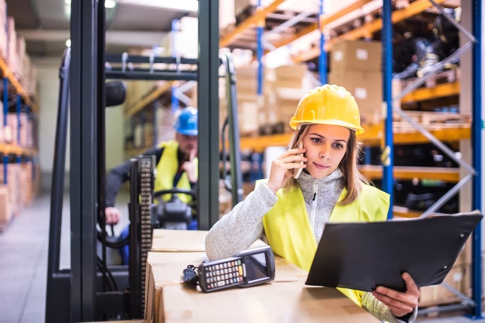 Female warehouse worker in safety vest using smartphone and tablet in distribution center